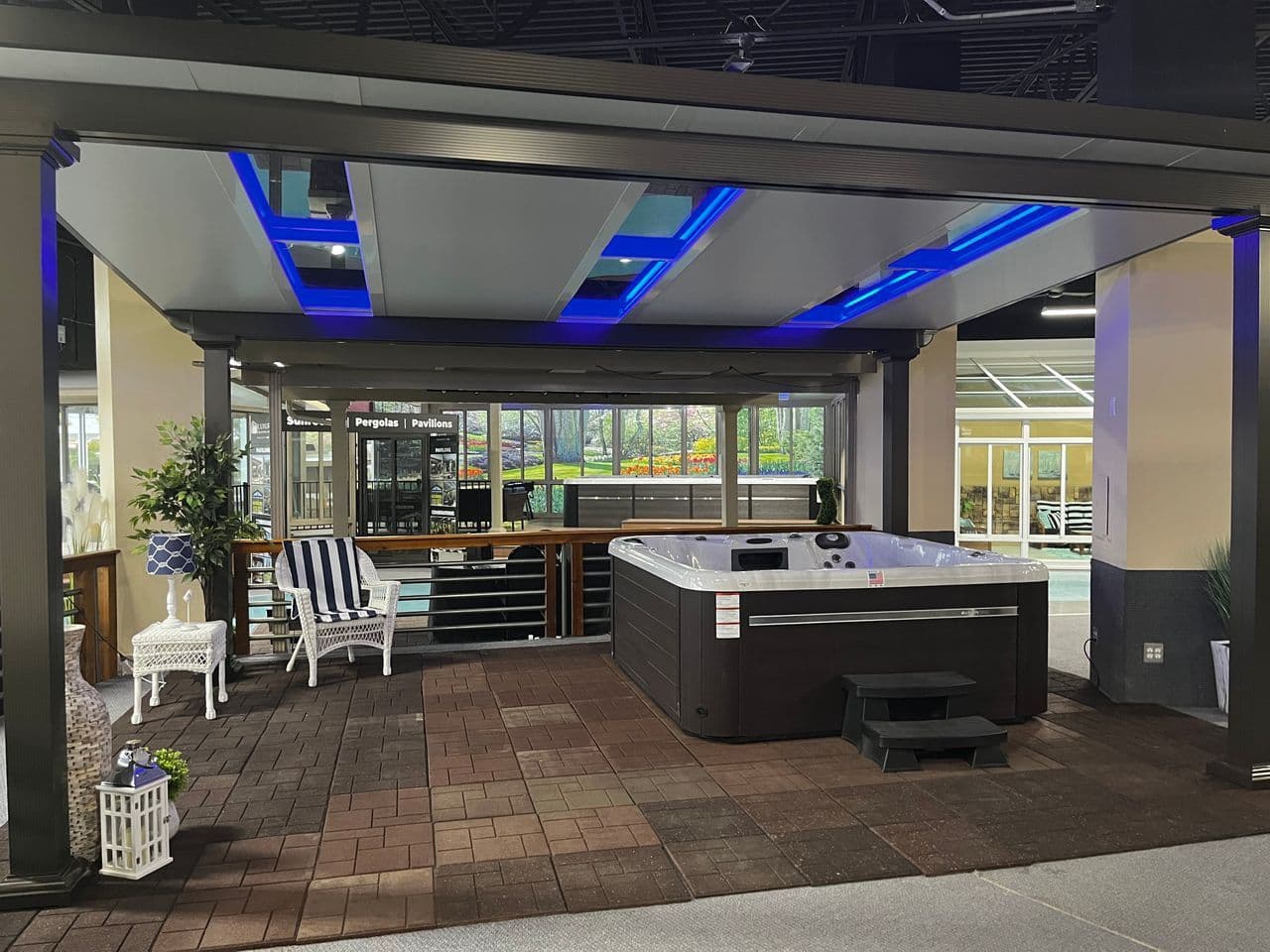A brick paved patio with a black pergola with skylights and blue LED lights. A hot tub is to the right side.